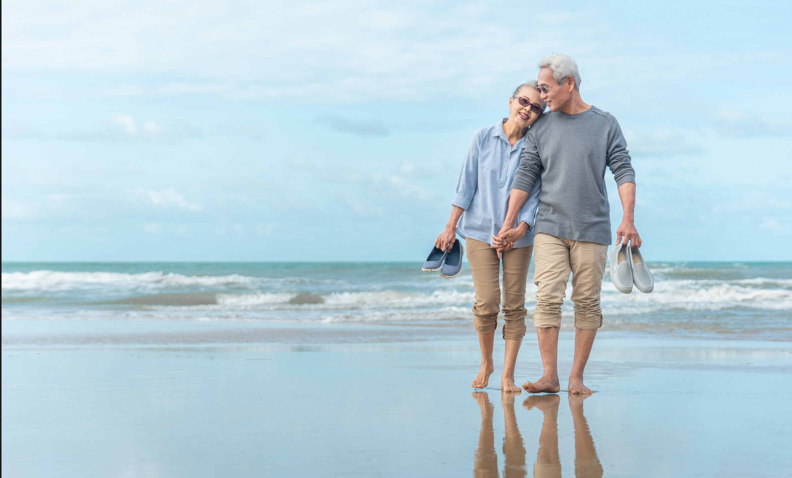 An older couple walks hand in hand along a calm beach, barefoot near the shoreline, holding their shoes and smiling at each other under a soft blue sky.